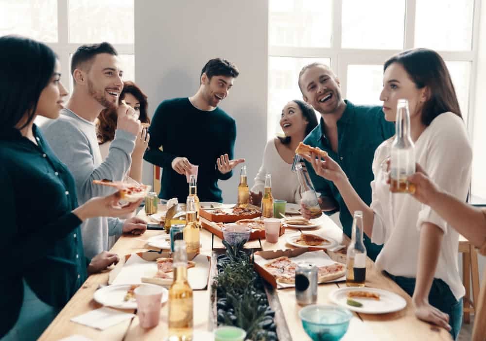 Group of young people in casual wear eating pizza and smiling while having a dinner party indoors